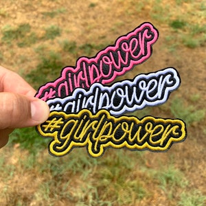 May include: Three embroidered patches with the text "#girlpower" in different colors. The top patch is pink, the middle patch is white, and the bottom patch is gold.