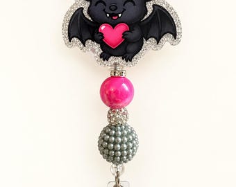 Puede incluir: Un portatarjetas con una figura de murciélago negro que sostiene un corazón rosa, rodeado por un borde de purpurina plateada. El portatarjetas está unido a una cadena de cuentas que incluye una esfera rosa grande, una cuenta de cristal pequeña y una esfera gris texturizada.