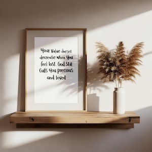 Può includere: Una stampa incorniciata con il testo "Your Value doesn't decrease when You feel lost. God Still Calls You precious and loved." è esposta su una mensola di legno. Un vaso bianco con piante essiccate è accanto. Lo sfondo è un muro neutro.