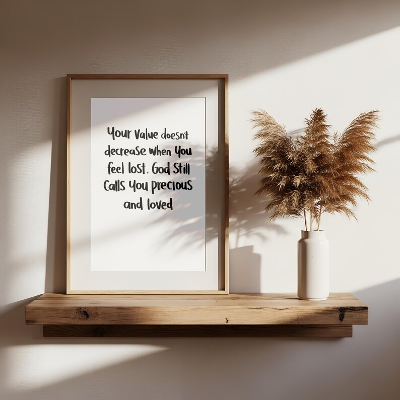 May include: A framed print with the text "Your Value doesn't decrease when You feel lost. God Still Calls You precious and loved." The print is displayed on a wooden shelf with a white vase holding dried plants. The background is a neutral wall.