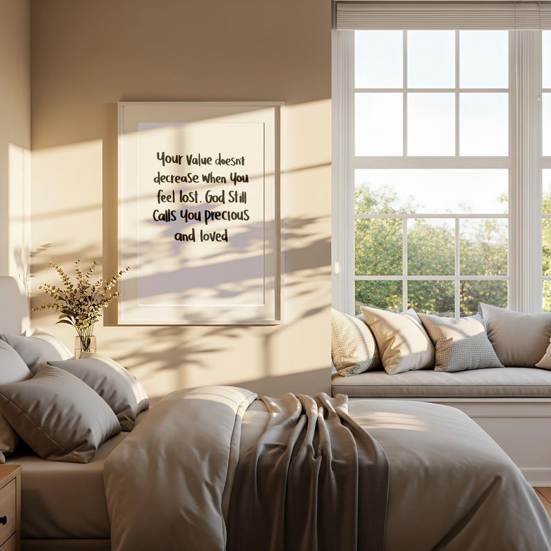 May include: A framed print with the text "Your Value doesn't decrease when you feel lost. God Still Calls You precious and loved" hangs above a bed with gray bedding. A vase of flowers sits on a nightstand.