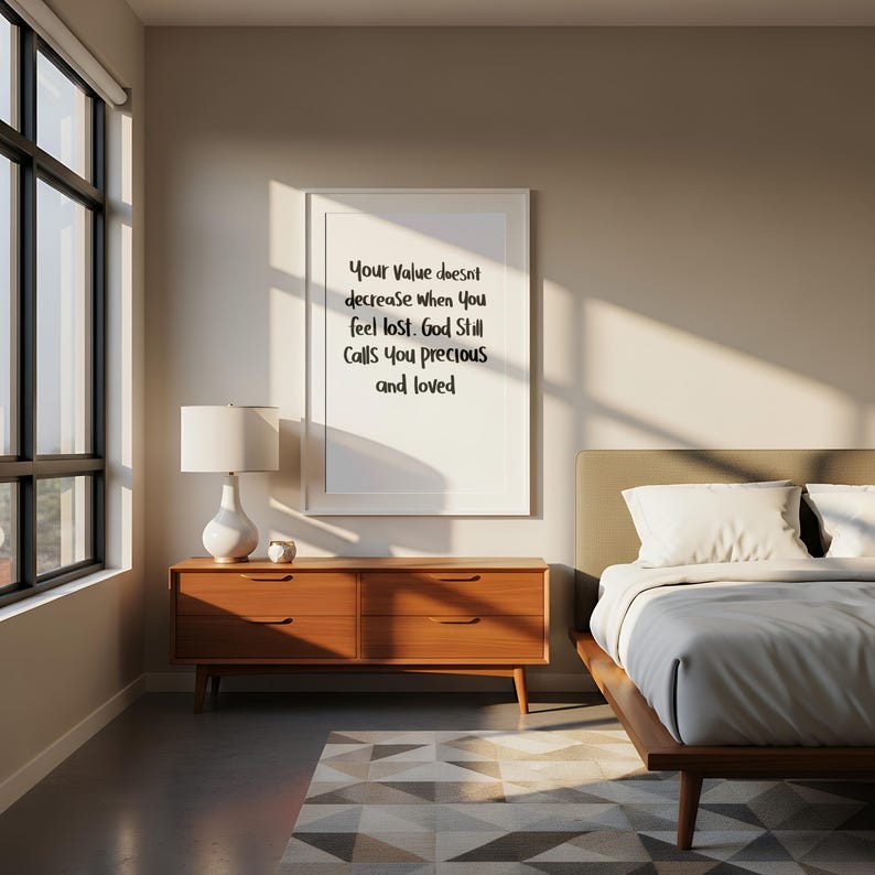 May include: A framed print with the text "Your Value doesn't decrease when You feel lost. God Still Calls you precious and loved" hangs above a wooden dresser. A white lamp sits on the dresser, and a bed is visible in the background.