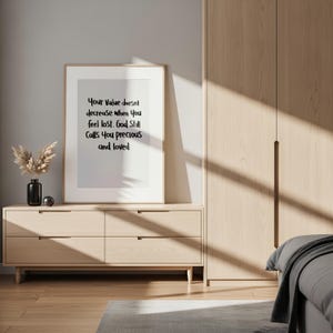May include: A framed print with the text "Your Value doesn't decrease when you feel lost. God Still Calls You precious and loved" sits on a light wood dresser. A black vase with dried flowers and a silver sphere are on the dresser.