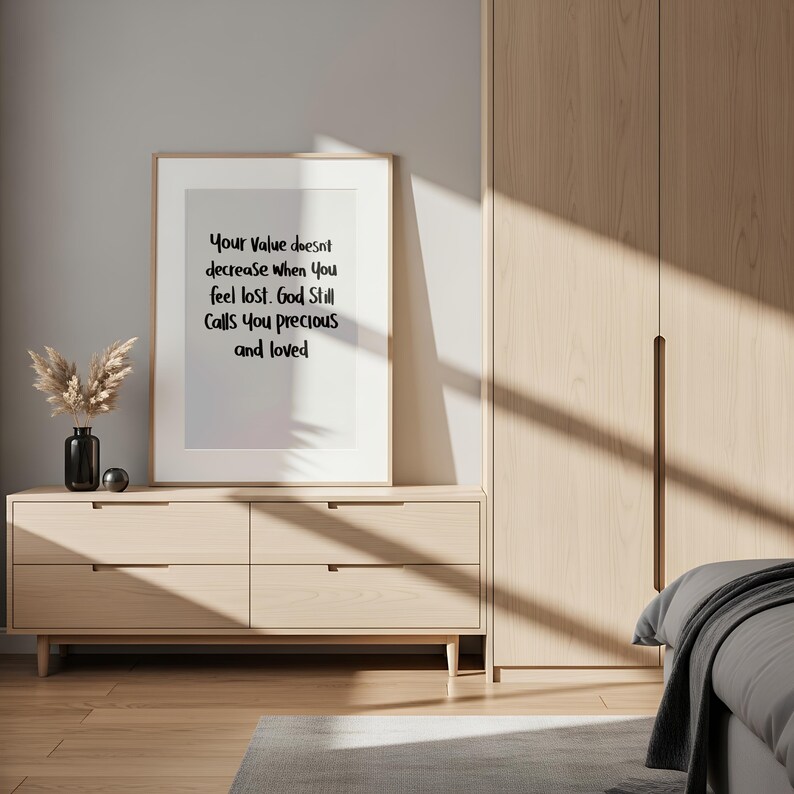 May include: A framed print with the text "Your Value doesn't decrease when you feel lost. God Still Calls You precious and loved" sits on a light wood dresser. A black vase with dried flowers and a silver sphere are on the dresser.