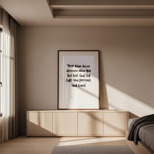May include: A framed print with the text "Your Value doesn't decrease when you feel lost. God Still Calls You precious and loved." The print is displayed on a light wood cabinet in a room with a window and curtains.