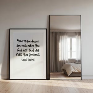 May include: A framed print with the text "Your Value doesn't decrease when You feel lost. God Still Calls You precious and loved" stands next to a tall, black-framed mirror. The mirror reflects a bedroom with a window and bed.