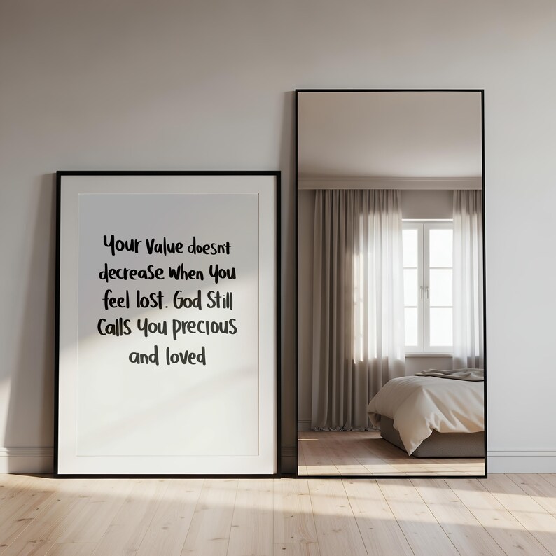 May include: A framed print with the text "Your Value doesn't decrease when You feel lost. God Still Calls You precious and loved" stands next to a tall, black-framed mirror. The mirror reflects a bedroom with a window and bed.