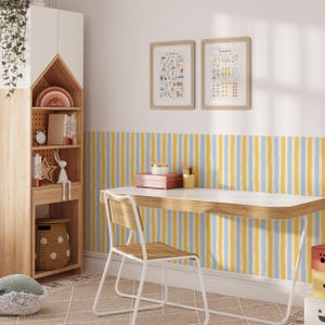 May include: A wooden house-shaped shelving unit with white doors, a desk with a white top and light wood trim, and a chair with a wooden seat and white metal frame. The wall features vertical yellow and blue stripes, with two framed educational posters.