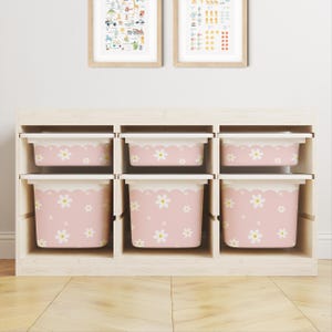 May include: A light-colored wooden storage unit featuring six removable plastic bins. Each bin is light pink with a white scalloped top edge and a repeating pattern of white daisy flowers with yellow centers. The unit has three columns and two rows.