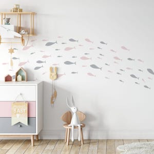 Può includere: Un muro bianco con una scuola di adesivi murali a forma di pesce rosa e grigi. Il muro è decorato con uno scaffale in legno con un aereo giocattolo e una stella in legno. Una cassettiera in legno con un cassetto rosa e grigio è in primo piano. Una sedia in legno con un giocattolo di coniglio bianco seduto su di essa è in primo piano. Il testo "sweet baby love" è su un cartello in legno appeso alla cassettiera.