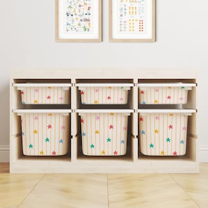 May include: A light-coloured wooden storage unit featuring six open compartments, each containing a plastic bin. The bins are decorated with a pattern of vertical yellow stripes and small, multi-coloured stars in pink, blue, and yellow.