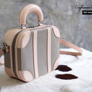 May include: A light pink leather handbag with a top handle and a shoulder strap. The bag has a gray canvas body with pink leather trim and a vintage suitcase design. The bag is sitting on a white and brown fur surface.