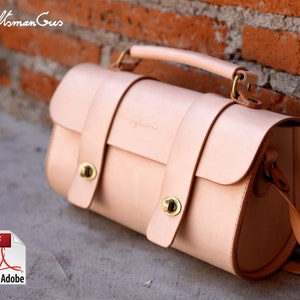 May include: A light tan leather handbag with a top handle and a shoulder strap. The bag has a rectangular shape with rounded edges and is secured with two straps and gold-coloured hardware. The text "Craftsman Gus" is visible on the bag.