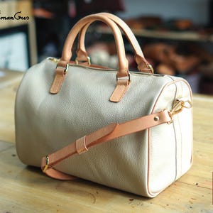 Puede incluir: Un bolso de mano de cuero granulado beige con dos asas superiores de color tostado claro y una correa ajustable a juego. El bolso tiene un cierre de cremallera superior con herrajes dorados y una etiqueta rectangular con el texto PDF Pattern.