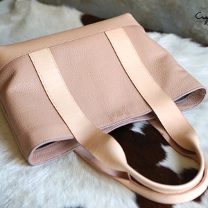 May include: A light pink leather tote bag with light tan straps. The bag has a textured surface and a rectangular shape. The bag is sitting on a white and brown fur-like surface. The text "Craftsman Gus PDF Pattern" is in the upper right corner.
