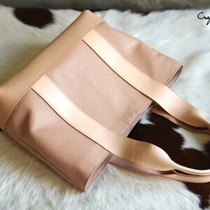 May include: A blush pink tote bag with light tan leather straps. The bag has a textured surface and a rectangular shape. The straps are attached to the top of the bag. The bag is sitting on a white and brown fur surface. The text "Craftsman Gus PDF Pattern" is in the upper right corner.