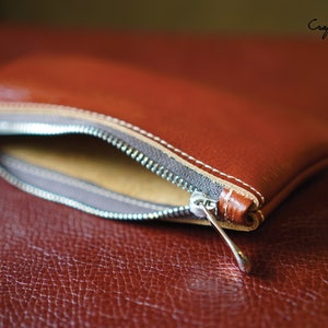 K&ouml;nnte beinhalten: Eine braune Leder-Rei&szlig;verschlusstasche mit einem silbernen Rei&szlig;verschluss-Zipper. Die Tasche ist offen und das Innere ist mit einem beigefarbenen Stoff ausgekleidet. Die Tasche liegt auf einer braunen Lederoberfl&auml;che. PDF-Muster.