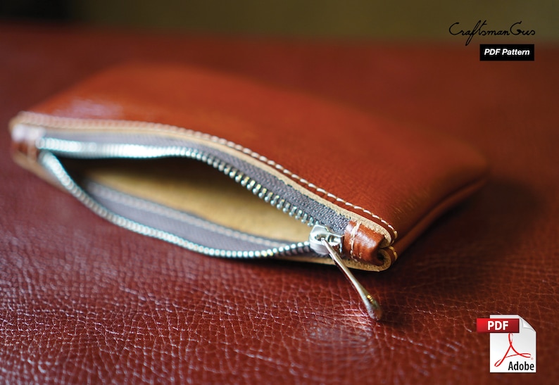 K&ouml;nnte beinhalten: Eine braune Leder-Rei&szlig;verschlusstasche mit einem silbernen Rei&szlig;verschluss-Zipper. Die Tasche ist offen und das Innere ist mit einem beigefarbenen Stoff ausgekleidet. Die Tasche liegt auf einer braunen Lederoberfl&auml;che. PDF-Muster.