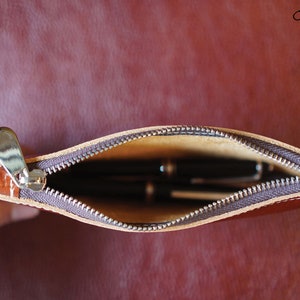 K&ouml;nnte beinhalten: Eine braune Leder-Rei&szlig;verschlusstasche mit einem Rei&szlig;verschluss-Zipper und einem beigefarbenen Futter. Die Tasche ist offen und enth&auml;lt mehrere Stifte.