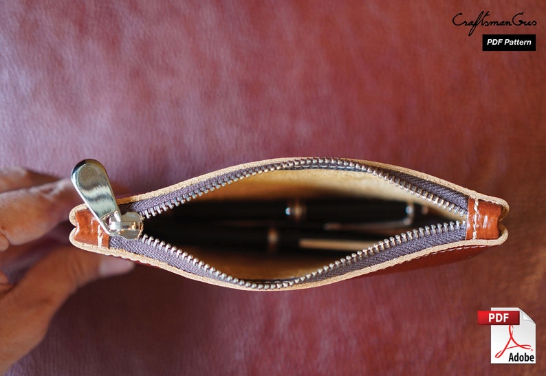 K&ouml;nnte beinhalten: Eine braune Leder-Rei&szlig;verschlusstasche mit einem Rei&szlig;verschluss-Zipper und einem beigefarbenen Futter. Die Tasche ist offen und enth&auml;lt mehrere Stifte.