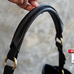 May include: Close-up of a black leather handbag handle with gold hardware. The handle is thick and padded for comfort.