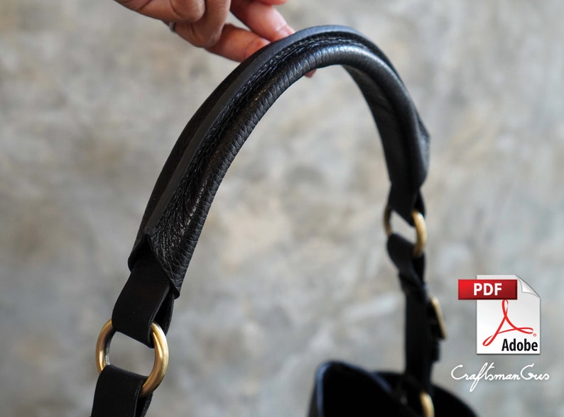 May include: Close-up of a black leather handbag handle with gold hardware. The handle is thick and padded for comfort.