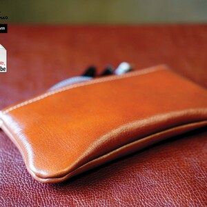 K&ouml;nnte beinhalten: Eine braune Leder-Rei&szlig;verschlusstasche oder -Geldb&ouml;rse auf einer roten, strukturierten Oberfl&auml;che. Die Tasche ist geschlossen und hat eine subtile W&ouml;lbung. Der Text "PDF Pattern" ist in der oberen linken Ecke des Bildes sichtbar.