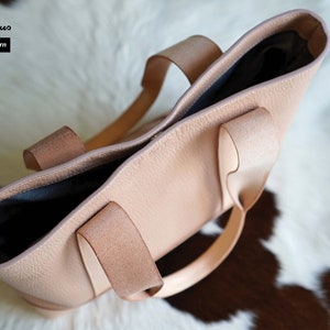 May include: A light pink leather tote bag with two handles. The bag has a textured surface and a dark interior. The text "Craftsman Gus PDF Pattern" is visible in the upper left corner. The bag is sitting on a white and brown surface.