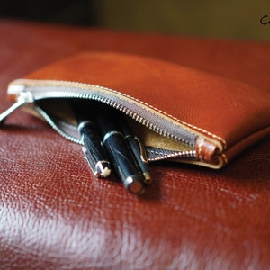 K&ouml;nnte beinhalten: Eine braune Leder-Rei&szlig;verschlusstasche mit zwei schwarzen Stiften darin. Die Tasche liegt auf einer braunen Lederoberfl&auml;che.  "Craftsman Gus PDF Pattern" steht in der oberen rechten Ecke des Bildes.