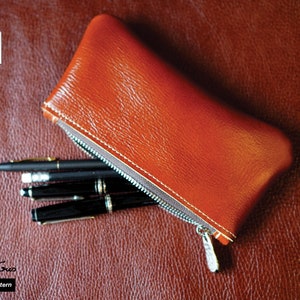 K&ouml;nnte beinhalten: Eine braune Leder-Rei&szlig;verschlusstasche mit einem Rei&szlig;verschluss-Zipper und vier schwarzen Stiften im Inneren. Die Tasche liegt auf einer braunen, strukturierten Oberfl&auml;che. Der Text "PDF Pattern" ist in der linken unteren Ecke des Bildes sichtbar.