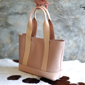 May include: A blush pink leather tote bag with light tan handles. The bag has a rectangular shape with a contrasting base. The handles are attached with vertical straps. The text "Craftsman Gus PDF Pattern" is visible.