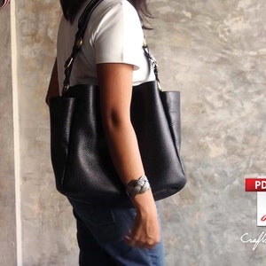 May include: A black leather tote bag with a braided strap and gold hardware. The bag is worn over the shoulder of a person wearing a white t-shirt and blue jeans.