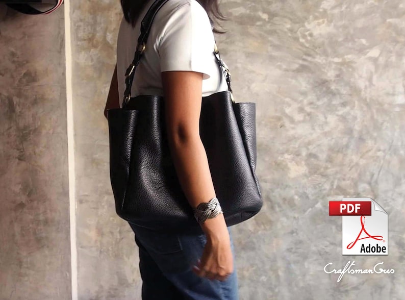 May include: A black leather tote bag with a braided strap and gold hardware. The bag is worn over the shoulder of a person wearing a white t-shirt and blue jeans.