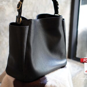May include: A black leather tote bag with a top handle and a buckle closure. The bag is sitting on a white and brown cowhide rug.