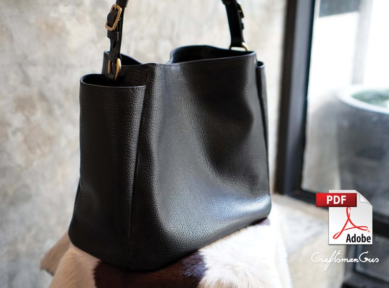 May include: A black leather tote bag with a top handle and a buckle closure. The bag is sitting on a white and brown cowhide rug.