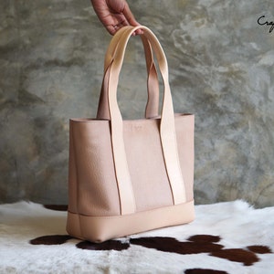 May include: A light pink leather tote bag with light beige straps and base. The bag has a rectangular shape and is held by a hand. The text "Craftsman Gus PDF Pattern" is visible in the upper right corner.