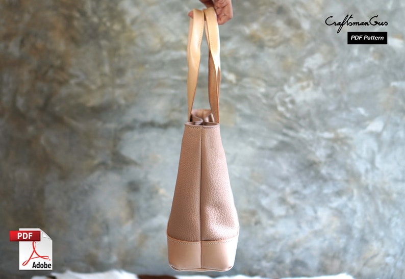 May include: A light brown tote bag with light tan handles and a light tan bottom. The bag has a textured surface and is held up by a person. The text "Craftsman Gus PDF Pattern" is in the upper right corner.