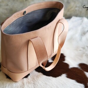 May include: A light brown leather tote bag with a textured finish. The bag has a rectangular shape with a flat bottom and two handles. The interior is a darker gray color. The bag is sitting on a white and brown cowhide rug. The text "Craftsman Gus PDF Pattern" is visible.