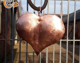 May include: A hammered copper heart ornament hanging from a metal hook. The heart is a deep reddish-brown color and has a shiny finish.