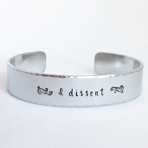May include: Silver bracelet with the text "I dissent" engraved on it. The bracelet has a hammered texture.
