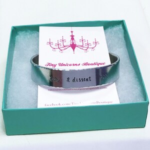 May include: A silver metal bracelet with the words "& I dissent" engraved on it. The bracelet is in a teal gift box with a white card that says "Tiny Unicorns Boutique".