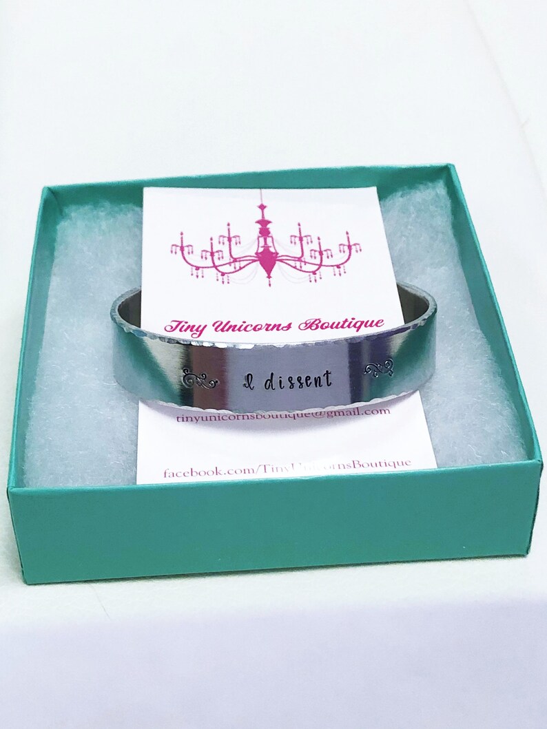 May include: A silver metal bracelet with the words "& I dissent" engraved on it. The bracelet is in a teal gift box with a white card that says "Tiny Unicorns Boutique".