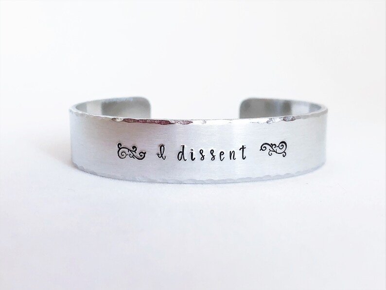 May include: A silver bracelet with the words "I dissent" engraved on it. The bracelet has a hammered finish and is decorated with two small swirls on either side of the text.