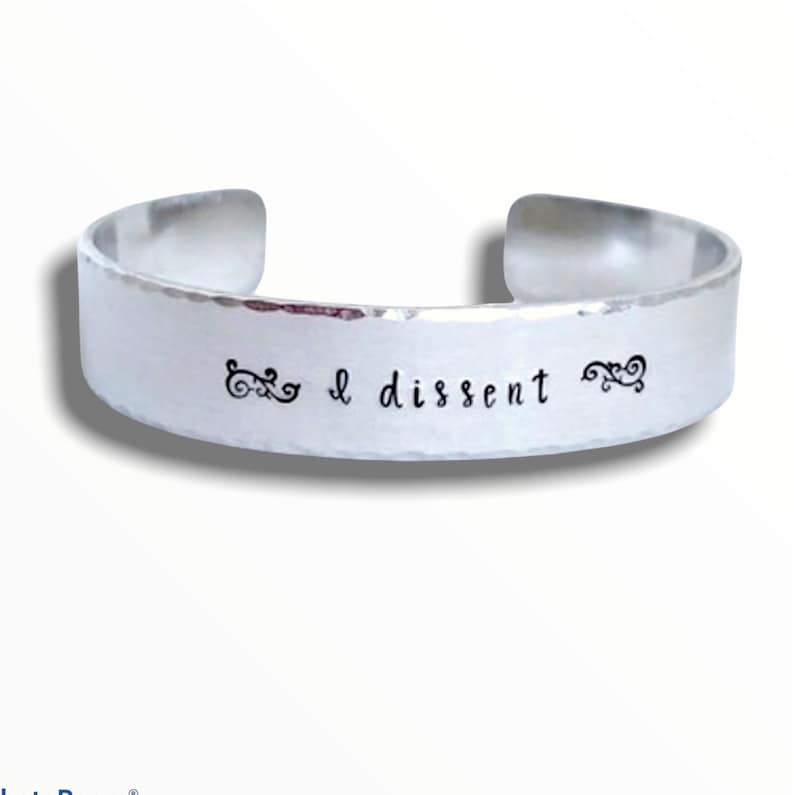 May include: A silver bracelet with the words "I dissent" engraved on it. The bracelet has a hammered finish and is decorated with two small swirls.