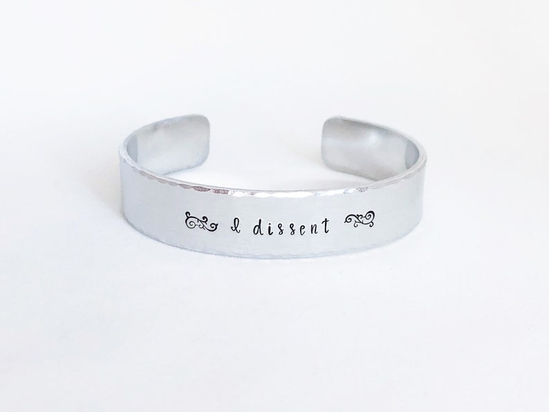 May include: A silver bracelet with the words "I dissent" engraved on it. The bracelet has a hammered finish and is decorated with two small swirls on either side of the text.
