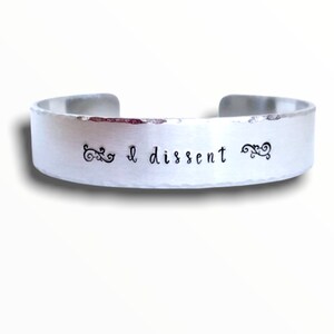May include: A silver metal bracelet with the words "I dissent" engraved on it. The bracelet has a hammered finish and is decorated with two small swirls on either side of the text.
