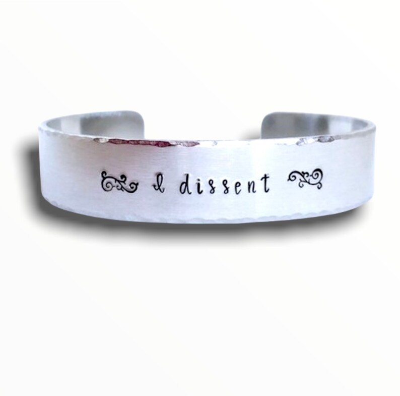 May include: A silver metal bracelet with the words "I dissent" engraved on it. The bracelet has a hammered finish and is decorated with two small swirls on either side of the text.