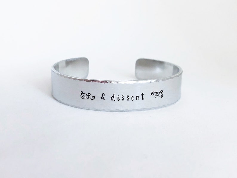 May include: Silver bracelet with the text "I dissent" engraved on it. The bracelet has a hammered texture.