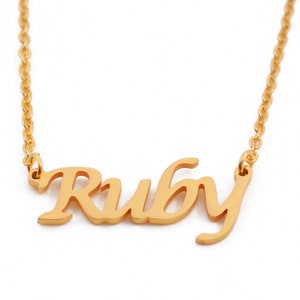 Può includere: Una collana color oro con il nome "Ruby" in corsivo. Il ciondolo è collegato a una catena delicata. La collana è mostrata su uno sfondo bianco.