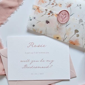 May include: A wedding invitation set featuring a floral patterned envelope sealed with a pink wax stamp. The invitation card reads "Rosie, will you be my Bridesmaid?" in pink script. A pink ribbon and envelope complete the set.